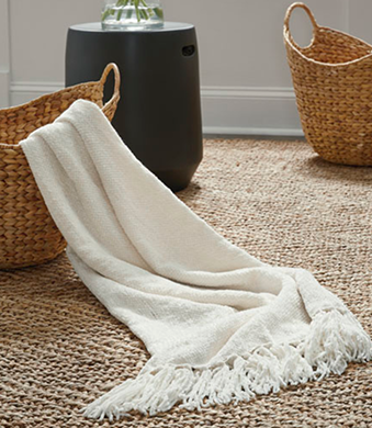 White throw blanket with fringe draped over a woven basket on a textured floor.